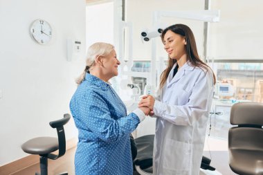 Mavi benekli tişörtlü yaşlı bir kadınla el ele tutuşan kadın doktor. İkisi de gülümsüyor ve iyi aydınlatılmış bir muayenehanede duruyor. Sağlık ve emekliler ve ilaç kavramı.