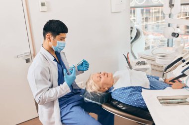 Yaşlı hasta sakin ve hazır görünürken dişçi ekipmanları hazırlıyor, modern klinik ortama güven ve bakım yansıtıyor. Sağlık ve emekliler ve ilaç kavramı.