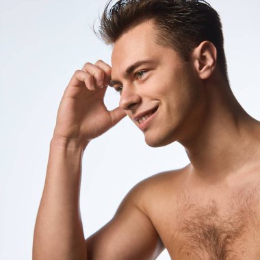 Yüzünün yanında el ele poz veren ve beyaz stüdyo arka planında kendine güvenen bir gülümsemeyle ileriye bakan üstsüz genç adamın yan görüntüsü. Bencillik ve sevgi kavramı, doğal güzellik, kozmetoloji.