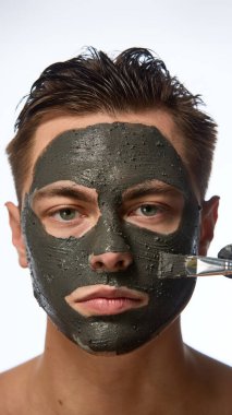 Pürüzsüz derili bir adam doğrudan öne bakıyor ve elinde fırça tutan siyah bir eldiven var. Yüzünün büyük bir kısmı beyaz arka planda maske takıyor. Bencillik, sevgi, güzellik, kozmetoloji kavramı.