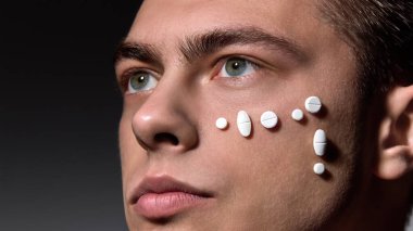 Man with smooth skin, white pills arranged in pattern on cheek, showing vitaminisation of skin, hair against dark studio background. Concept of selfcare and love, natural beauty, cosmetology. clipart