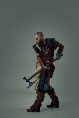 Çift başlı baltayı kavrayan Viking savaşçısı, savaşa hazır bir vaziyette çömelmiş, kürk pelerin giyiyor ve karanlık stüdyo geçmişine karşı dayanıklı zırh giyiyor. Ortaçağ savaş alanı kavramı, savaşçı ruhu..