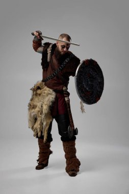 Kürk pelerinli ve deri zırhlı Viking savaşçısı gibi giyinmiş bir adam stüdyo arka planına kılıç darbesi hazırlarken İskandinav gravürlü bir zırh tutuyor. İskandinav kültürü kavramı, Viking mirası