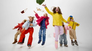 Bir grup çocuk, havada, şapkalarını çıkarıyorlar, rahat kışlık ceketler giyiyorlar, eğlence ve kahkahaları beyaz stüdyonun arka planında kutluyorlar. Çocukluk kavramı, kış tatili, tasasız.