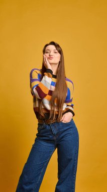 Genç bir kadın, neşeli bir ifadeyle, el ele tutuşmuş, öpüşüyor, çizgili süveter giyiyor, hardal rengi stüdyo arka planına karşı. Flört kavramı, neşe, gençlik, aşk.