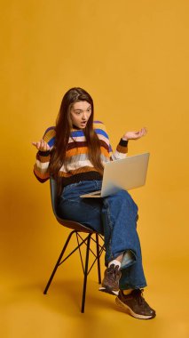 Genç bir kadın, kafası karışmış, dizüstü bilgisayarla oturuyor, hayal kırıklığı içinde ellerini kaldırıyor, çizgili kazak giyiyor, hardal rengi stüdyo geçmişine karşı. Teknoloji kavramı, online alışveriş.