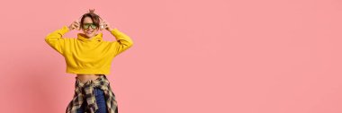 Genç bir kadın, neşeli bir ifadeyle, güneş gözlüğü takıyor, sarı kapüşonlu ve kareli gömlek giyiyor, pembe stüdyo geçmişine karşı. Gençlik kavramı, güven, kentsel yaşam tarzı, müzik kültürü.