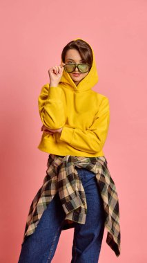Kendine güvenen, güneş gözlüğü takan, sarı kapüşonlu ve kareli tişörtlü pembe stüdyo geçmişli genç bir kadın. Tavır kavramı, eğilim, bireysellik, kendini ifade etme.