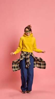 Genç bir kadın, kaygısız bir ifadeyle, hareket ederken kareli gömleğini çeviriyor, sarı kapüşonlu, pembe stüdyo geçmişine karşı. Eğlence, enerji, dinamik yaşam tarzı, moda kavramı.