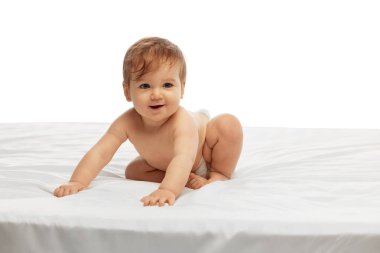 Küçük bebek, beyaz arka planda merak ve heyecanla ileriyi seyrederken yumuşak beyaz yatakta emekliyor. Çocukluk kavramı, gelişme, doğma büyüme, hareket, öğrenme.