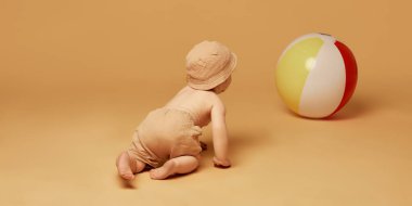 Meraklı bir bebeğin geniş renkli bir plaj topuna doğru süründüğünü yumuşak şapkalı ve şortlu sıcak bej arka planda. Keşif kavramı, hareket, keşif, çocukluk sevinci.