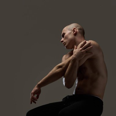 Adam oturmuş, kollarını göğsüne dolamış, gözleri yoğun, koyu kahverengi arka plana karşı kontrol ve hassasiyet yayıyor. Cesur varlık kavramı, yontulmuş zarafet, güç, erkeklik..