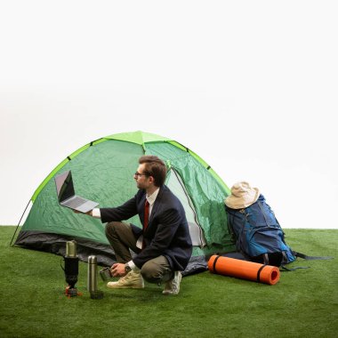 İş adamı elinde dizüstü bilgisayarla kamp çadırının yanında çömelmiş beyaz stüdyo arka planına termostan su döküyor. Mobil ofis kavramı, dijital göçebe, iş hayatı entegrasyonu, bağımsızlık.