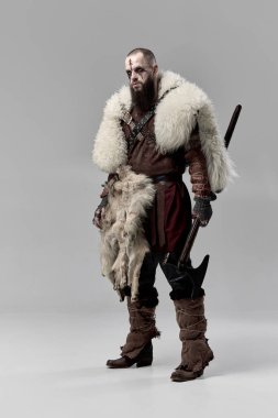 Kararlı bir adamın tam boy portresi, deri zırh ve kürk pelerinli ortaçağ Viking 'i, gri stüdyo arka planına karşı baltayla ayakta duruyor. Tarihi portre kavramı, İskandinav mitolojisi
