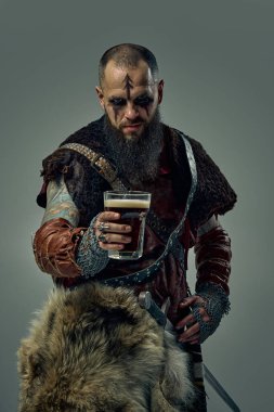 Kürk pelerin ve deri zırh giymiş bir ortaçağ Viking savaşçısının portresi. Elinde kılıç, elinde bir bardak bira, stüdyo arka planında duruyor. İskandinav gelenekleri kavramı, bira festivali