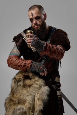 Altın bira şişesini kavramış Viking savaşçısının portresi, kürk pelerin ve stüdyo arka planında giyilmiş deri zırh. İskandinav ziyafeti, ortaçağ savaşçı kültürü, İskandinav kutlaması.