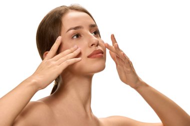 Genç, güzel bir kadın parmak uçlarıyla nazikçe masaj yapıyor ve beyaz stüdyo arka planına bakıyor. Model rahat görünüyor. Cilt bakımı rutini, rahatlama, yüz masajı, sağlık..