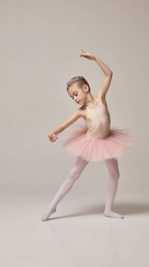 Zarif bir çocuk, sevimli küçük balerin, pembe etek ve taç giymiş, sıcak gri arka planda dans hareketleri yapıyor. Bale anlayışı, zarafet ve güzellik, zarafet. Ad