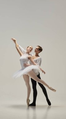 Klasik pozda balerin kolunu uzatıyor, dengeli duruş içinde partnerin arkasında duruyor, beyaz stüdyo arka planına karşı tutu hareketini arttırıyor. Klasik bale anlayışı, saflık, duruş, güzellik.