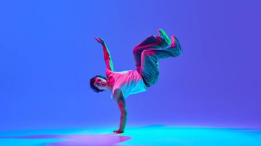 Enerjik genç adam break dans yapıyor, neon ışıkta parlak mavi-mor arka plana karşı güç ve çeviklik gösteriyor. Sokak dansı, hareket, tutku, gençlik kültürü ve yaratıcılık kavramı.