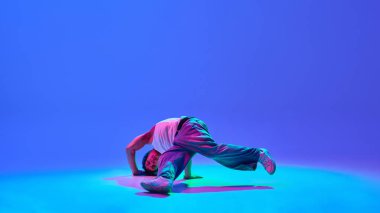 Hareket halindeki atletik genç adam, neon mavisi arkaplana karşı havadaki break dans dönüşü, enerji ve yaratıcılığı ifade ediyor. Sokak tarzı, hip-hop hareketi ve şehir kültürü kavramı.