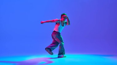 Erkek performansının dinamik görüntüsü, neon ışıkta değişen mavi arkaplana karşı günlük kıyafetlerle break dans. Dans tutkusu, enerji ve serbest sanatçılık kavramı.