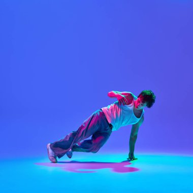 Erkek performansının dinamik görüntüsü, neon ışıkta değişen mavi arkaplana karşı günlük kıyafetlerle break dans. Dans tutkusu, enerji ve serbest sanatçılık kavramı.