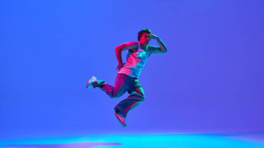 Yetenekli ve yetenekli genç adam hareketli gündelik giysiler içinde, neon ışıkta mavi renkli arka planda dans ediyor. Break dans kavramı, hip-hop hareketi, sokak performansı