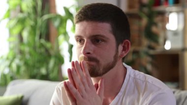 Genç sakallı, beyaz tişörtlü, oturma odasında kanepede oturan endişeli ve gergin görünen bir adam. İnsan duyguları, stres, zorluklar, akıl sağlığı kavramı. Ad