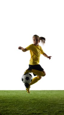 Kızın dikey görüntüsü, sarı formalı futbolcu ve siyah şortlu, topla hareket eden, beyaz stüdyo arka planına karşı atlayan. Spor, çocukluk, hareket, rekabet, etkinlik kavramı