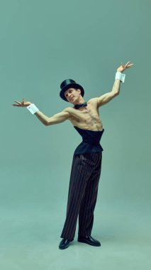 Yapılandırılmış korse ve ince çizgili pantolonlar içinde sessiz yeşil stüdyo arka planına karşı hareketlerle poz veren etkileyici bir sanatçı. Kabare estetiği, klasik enerji, dans, cazibe kavramı.