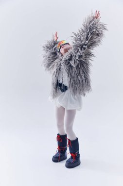 Büyük boy kürk manto, kayak gözlükleri ve ay botlarıyla beyaz stüdyo arka planında dans eden genç bir kadın. Şen şakrak zarafet kavramı, kış eğilimleri, sanatsal ifade, lüks kış tarzı.