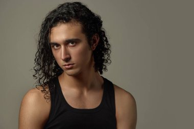 Uzun, kıvırcık saçlı ve pürüzsüz tenli özgüvenli genç adamın portresi tarafsız stüdyo arka planına yoğun bir şekilde kameraya bakıyor. Erkek güzelliği, moda, cilt bakımı, kozmetoloji, kozmetik.