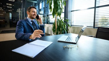 Telefon görüşmesi sırasında gülümseyen neşeli işadamı, modern geometrik tavan lambaları altında dizüstü bilgisayar ve ofis malzemeleriyle masa başında. Ağ kurma, kurumsal iletişim, liderlik kavramı.