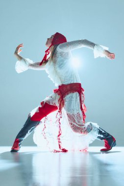 Dalgalı dantelli ve kırmızı dokuları olan genç bir kadın vücudunu esnetiyor, kolları manevi hareketlerle kıvrılmış, ruhani arka plan ışığıyla aydınlatılıyor. Sürrealizm kavramı, çağdaş performans, moda, hareket.