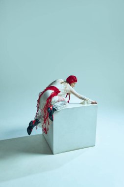 Beyaz küpün üzerinde sürünen süslü kıyafetli genç bir kadın, parmakları uzamış, botları havada, mücadele ve dönüşümü çağrıştırıyor. Moda sürrealizmi kavramı, hareket psikolojisi, rüya gibi..