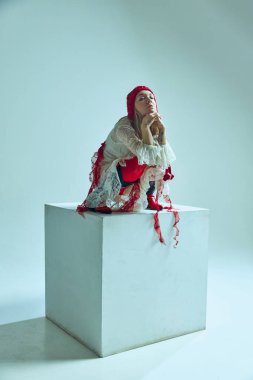 Beyaz dantelli elbise içinde beyaz küpün üzerine tünemiş genç bir kadın, kırmızı tayt, dalgın bir poz. İç gözlem kavramı, moda sürrealizmi, modern folklor, duygusal derinlik, sanatsal hareket.