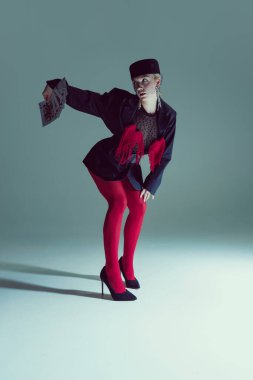Siyah ceketli genç bir kadın, kırmızı tayt ve kartları uzatırken çömelmiş topuklu ayakkabılar. Moda, strateji, gizem, şans oyunu, avangard performans, görsel sembolizm kavramı.
