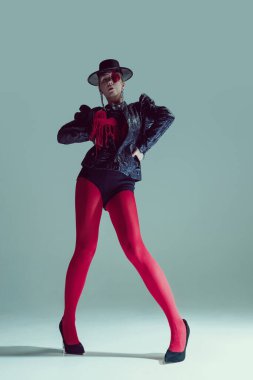 Kırmızı taytlı ve gotik ceketli kadın baskın bir duruş sergiliyor, kalp kapaklı gözleriyle bakıyor. Moda ve tarz kavramı, gotik romantizm, gizem, psikolojik derinlik.