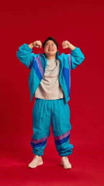 Asyalı genç bir adamın eski spor kıyafetlerini giymiş ve kırmızı arka plana karşı gücünü ve güvenini gösteren uzun bir fotoğrafı. İnsani duygular, sağlık, enerji kavramı. Ad