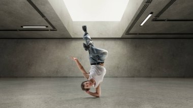 Gülen dansçı başıyla dengede duruyor, bacağı kalkık, neşeli hip-hop pozunda donmuş, yumuşak ışık altında gri beton duvarlara karşı. Özgürlük kavramı, gençlik, ritim, dans eden ruh beton şehir çerçevesinde