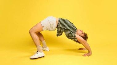 Genç adam atletizm kıyafeti giymiş, esneklik egzersizleri yapıyor, parlak sarı stüdyo geçmişine karşı köprü. Spor egzersizleri, aerobik, dayanıklılık, fiziksel sağlık ve refah kavramı.