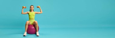 Pankart. Çekici, atletik bir kız, fitness topunun üzerinde oturuyor ve dambıllarıyla fotokopi çekilen mavi arka plana karşı eller için egzersiz yapıyor. Spor egzersizleri, sağlık, fiziksel sağlık kavramı.