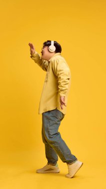 Genç Asyalı bir adam, şık giyinmiş günlük kıyafetler ve gözlükler takmış, kulaklıkla güneşli sarı arka planda müzik dinlerken dans ediyor. İnsan duyguları, moda, tarz, kendini ifade etme kavramı. Ad