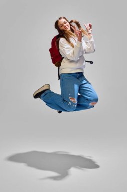 Sırt çantalı kız heyecanla hareket ediyor, barış işareti gösteriyor, okul gününün neşesini ve özgürlüğünü simgeliyor. Eğitim, tatiller, olumlu duygular, eğlence ve neşe kavramı.