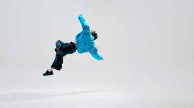 Genç dansçı havada kıvranıyor, bacakları dinamik bir şekilde beyaz stüdyo arka planına doğru uzanıyor. Kontrollü hareket. Dans kültürü kavramı, aktif yaşam tarzı, güç, ritim, performans.