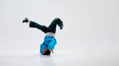 Dinamik sokak dansçısı, eski moda giyinmiş, kafası üzerinde dönüyor, bacakları beyaz arka plana doğru uzanıyor. Müzik ve hareket kavramı, kendini ifade etme, enerji, modern sanatçılık.