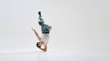 Breakdansçı, günlük giysiler giymiş, tek elle donduran, bacaklarıyla beyaz arka plana uzanan biri. Müzik kavramı, şehir hareketi, beceri, enerji, modern sanat.