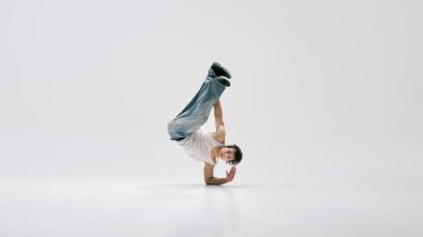 Yetenekli hip hop dansçısı, günlük giysiler giymiş, vücudu ters bir pozisyona sokup beyaz arka plana karşı tam kontrol ve dinamik duruş sergilemiş. Güç kavramı, dayanıklılık, break dans, hareket.