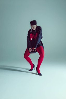 Kırmızı detayları olan koyu siyah ceketli genç bir kadın, kırmızı taytlı, beyaz arka plana karşı dinamik bir duruşla çömelmiş. Moda ve tarz kavramı, kadınlık, psikoloji, kendini ifade etme.